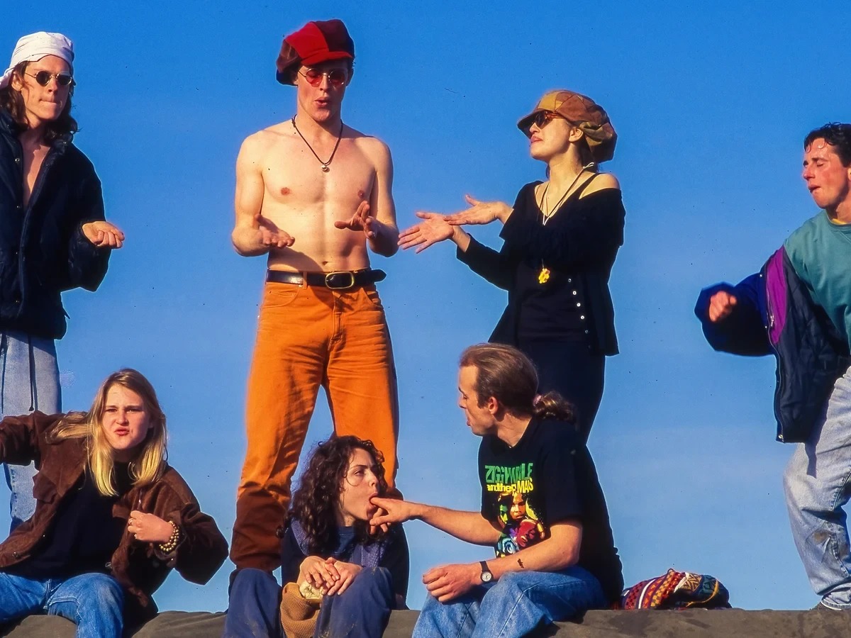 Seven festivalgoers on a ridge at Castlemorton against a clear blue sky in 1992; four of them stand with their hands raised and turned over, none of them looking at each other, while on the ledge below a young woman in round glasses holds the index finger of the man sitting beside her in her mouth and nobody in the frame reacts