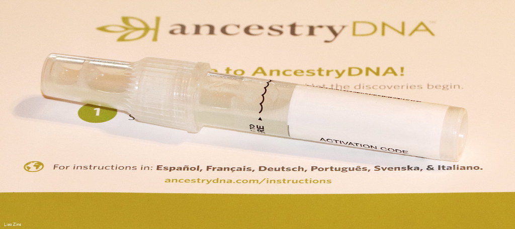 A clear plastic saliva collection tube, half full, lying on the printed instruction card from a mail-order ancestry DNA kit; the card reads ancestryDNA and let the discoveries begin, and the tube carries a label reading ACTIVATION CODE